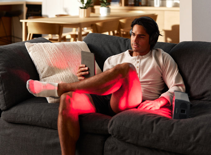 Man sitting on couch with headphones, with a red light device on the couch next to him shining on his leg.